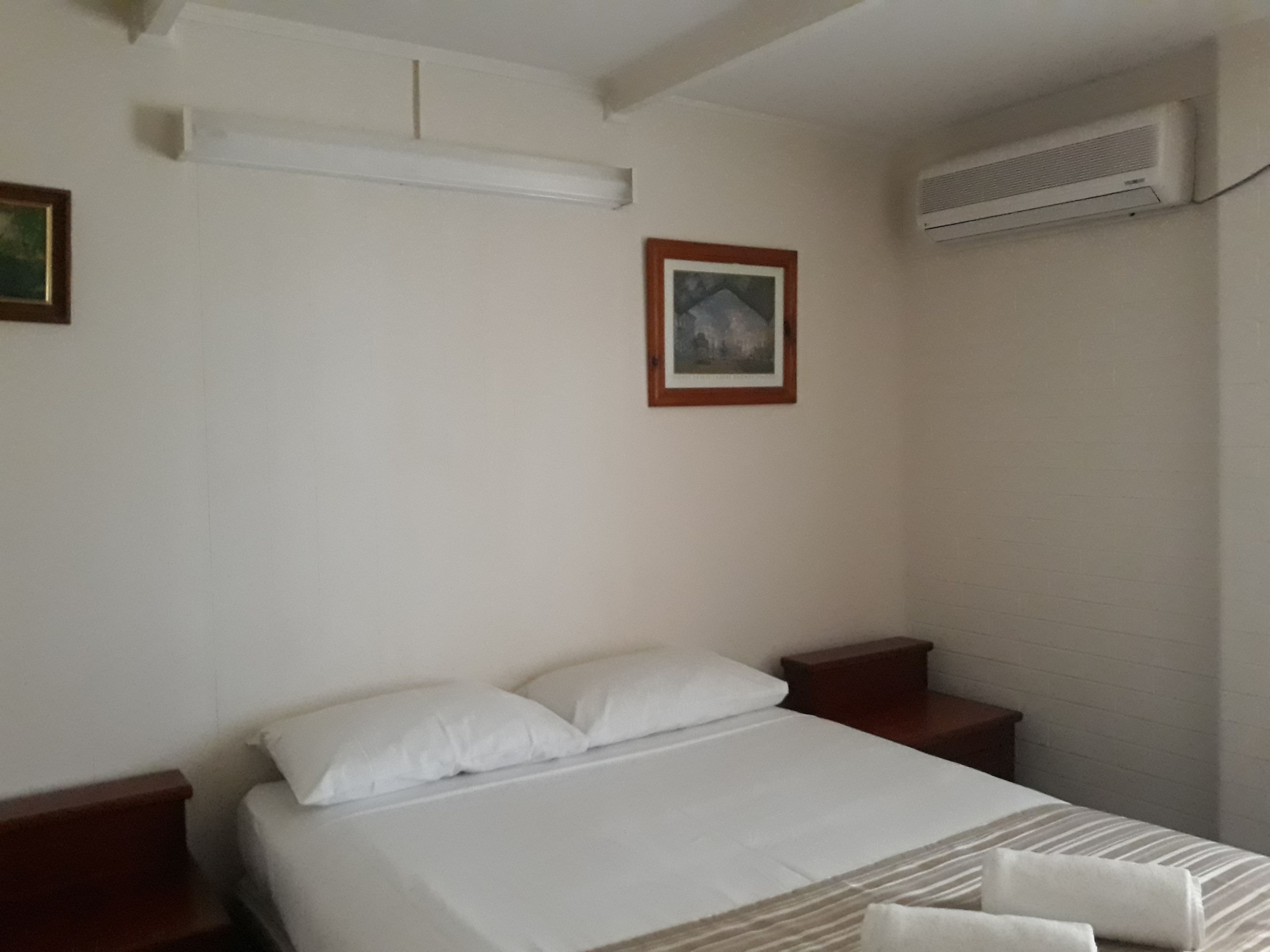A hotel room with a single bed, a nightstand, a framed picture on the wall, and an air conditioning unit.