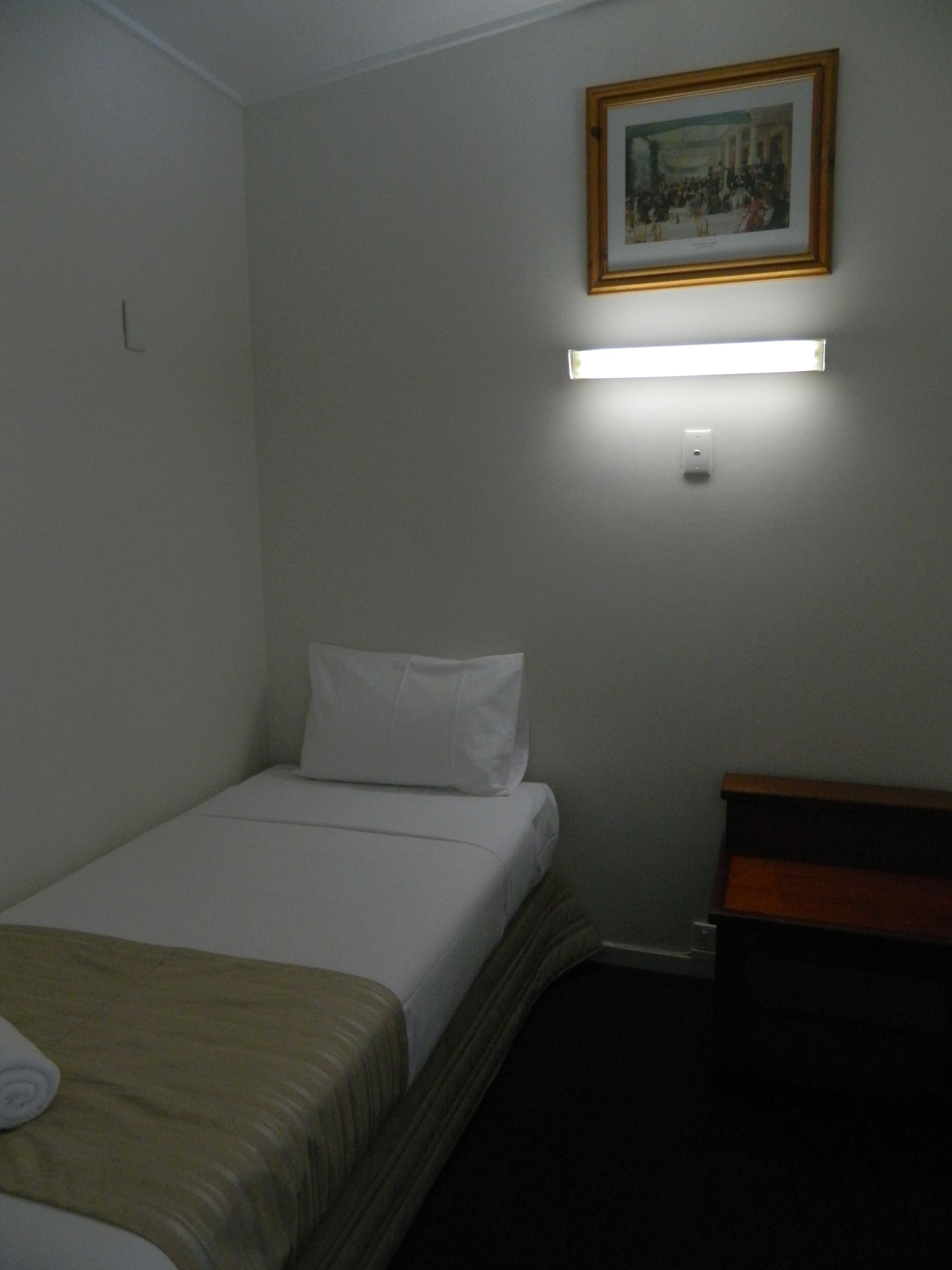 Una cama individual con ropa de cama blanca, una mesita de noche de madera, una lámpara fijada a la pared y un cuadro enmarcado sobre la cama.