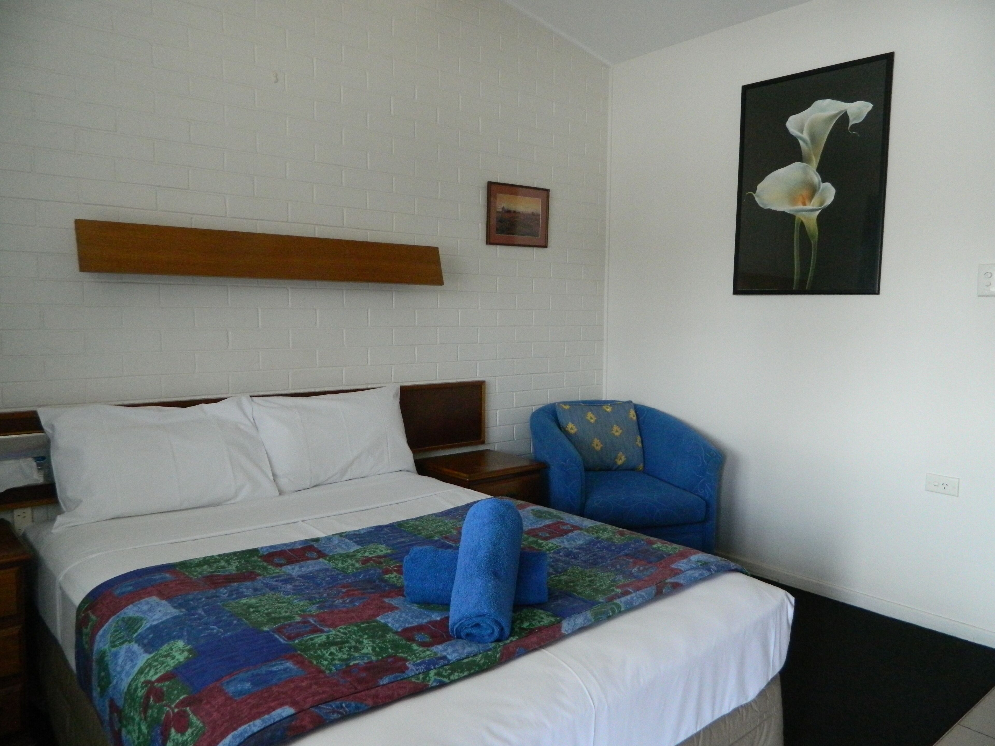 Habitación de hotel con una cama, un sillón azul, una colcha y un cuadro de una flor colgado en la pared.