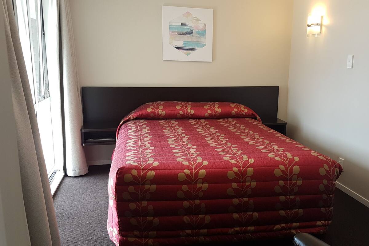 A hotel room with a bed, a nightstand, a wall-mounted light, and a window with curtains.