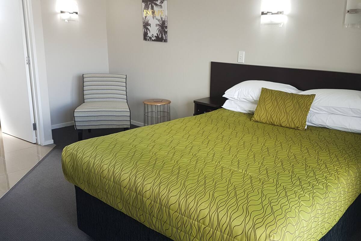 A hotel room with a large bed, a chair, a small table, and a wall-mounted light fixture.