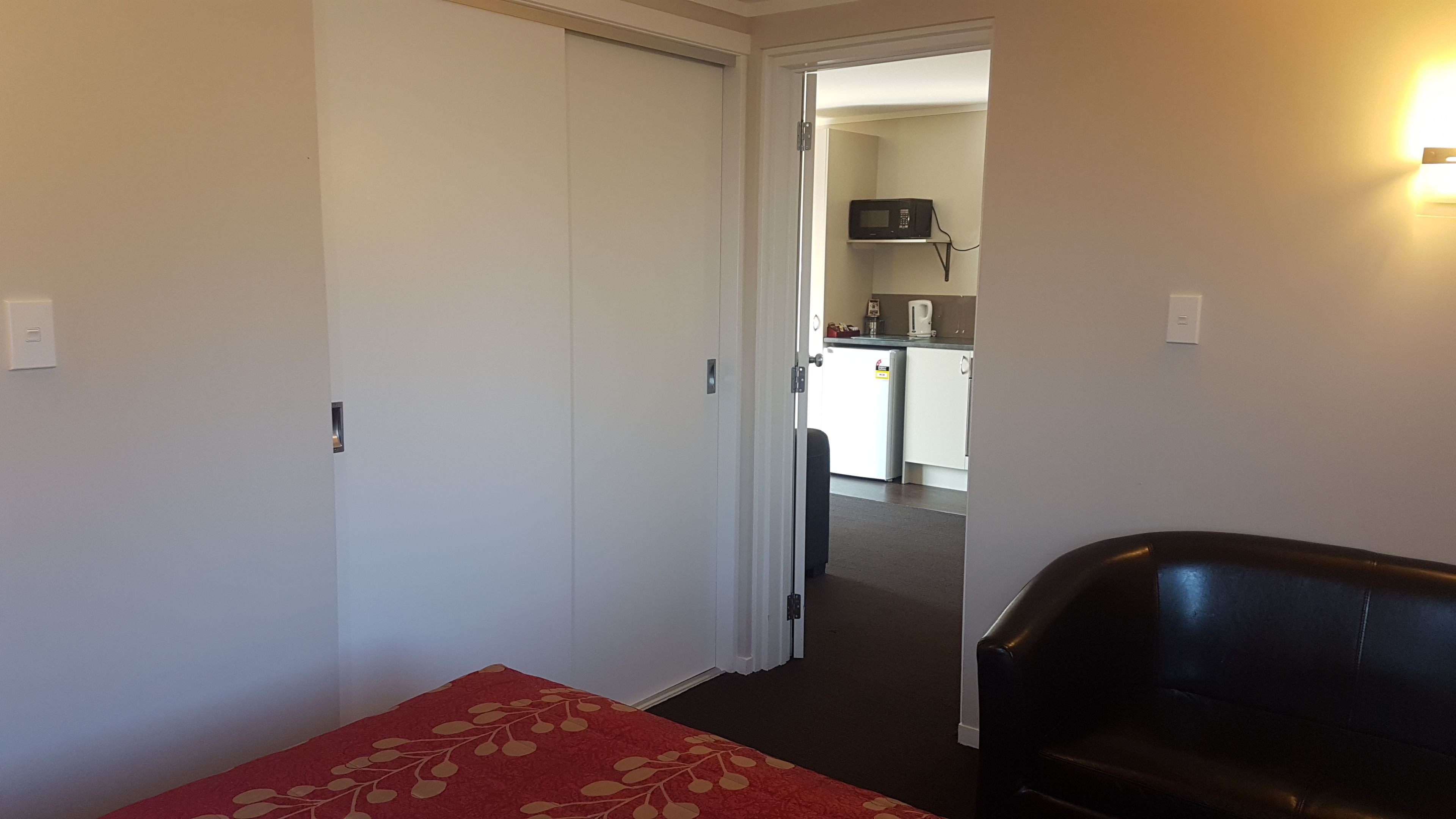 Ein Hotelzimmer mit einem Bett, einer roten blumigen Tagesdecke, einem schwarzen Ledersessel und einer Küchenzeile im Hintergrund.