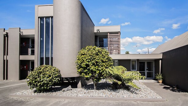 A modern building with a curved facade, large windows, and a small garden area with shrubs and a potted plant. - Aspree Motor Inn (Palmerston North)