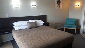 A neatly made bed with a patterned bedspread, a blue chair, and two wall-mounted lights. - Aspree Motor Inn (Palmerston North)