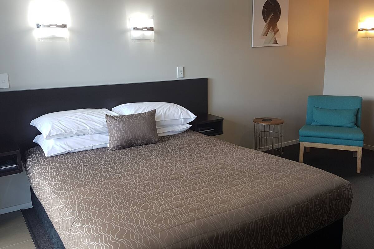 A neatly made bed with a patterned bedspread, a blue chair, and two wall-mounted lights.