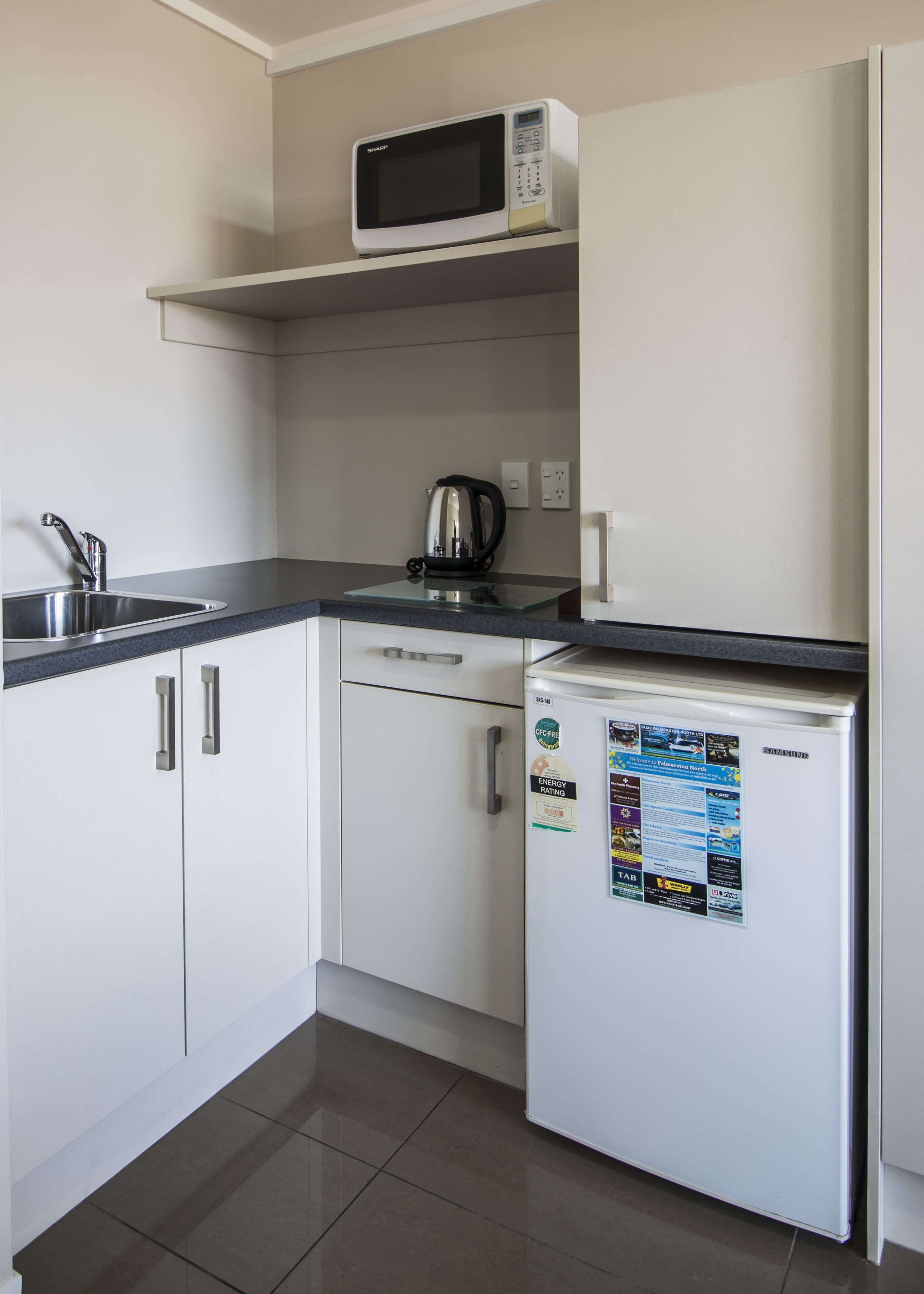 Een compacte keuken met witte kastjes, een roestvrijstalen spoelbak, een magnetron, een waterkoker en een koelkast met energielabels.