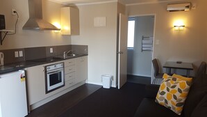 A compact kitchen with a stove, sink, and cabinets. There is a small trash bin and a dining area with a table and chairs. - Aspree Motor Inn (Palmerston North)