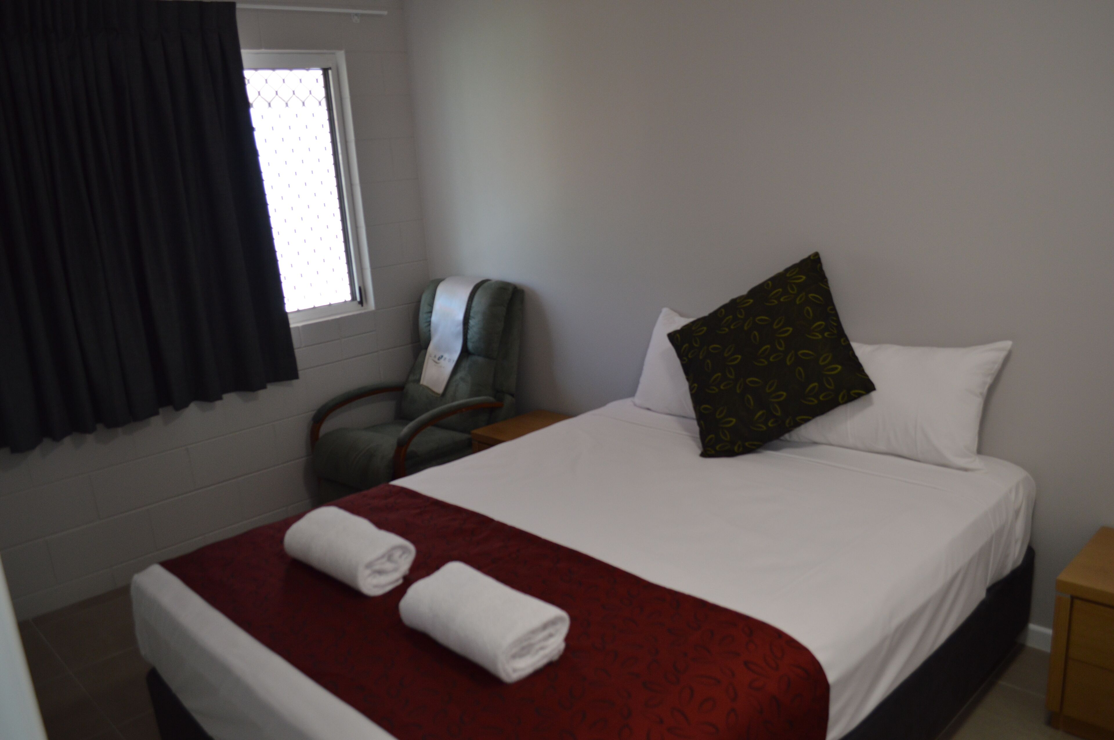 A hotel room with a neatly made bed, a chair, a small table, and a window with curtains.