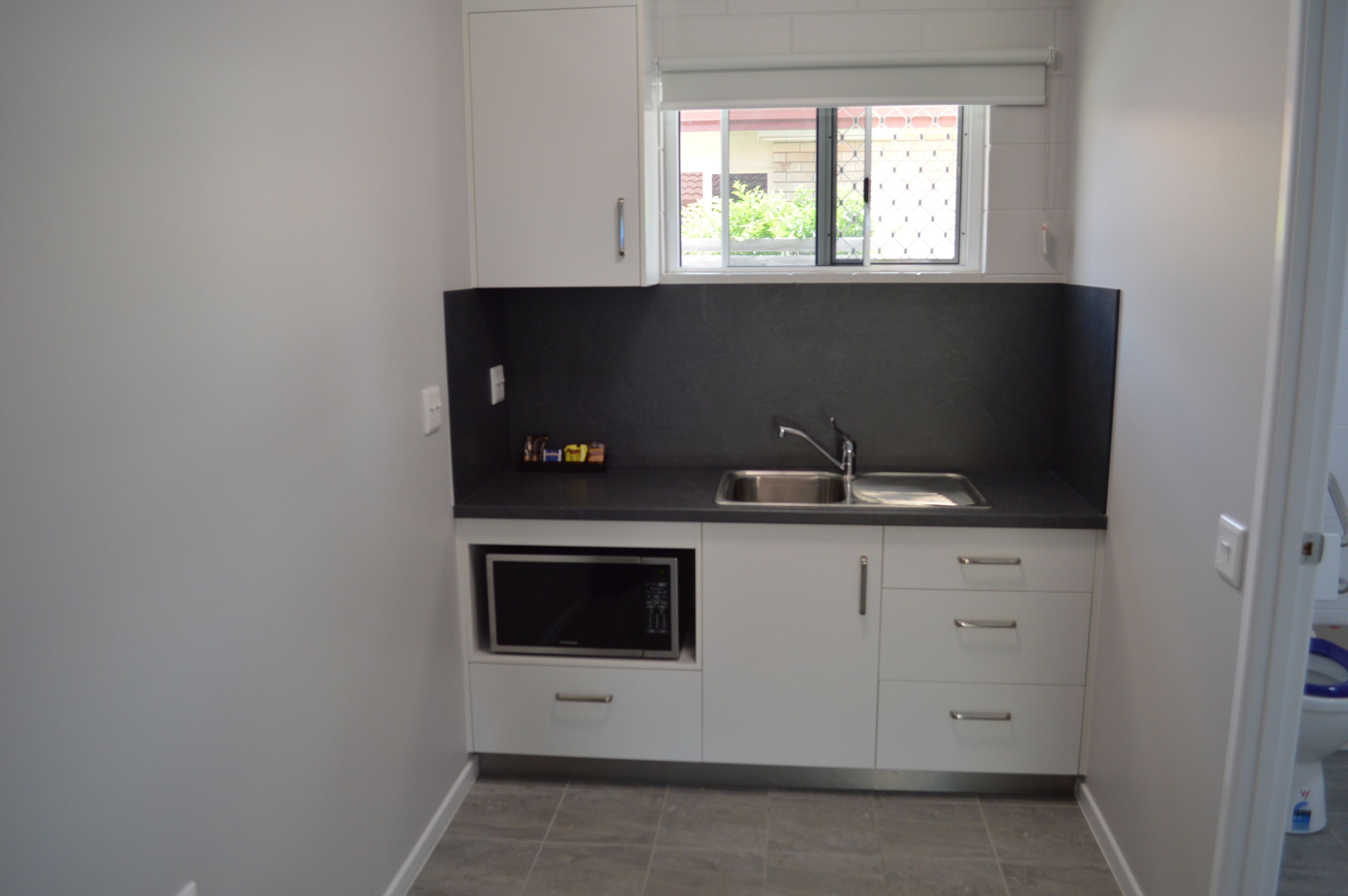 A compact kitchen with white cabinets, a black countertop, a built-in microwave, and a window above the sink.
