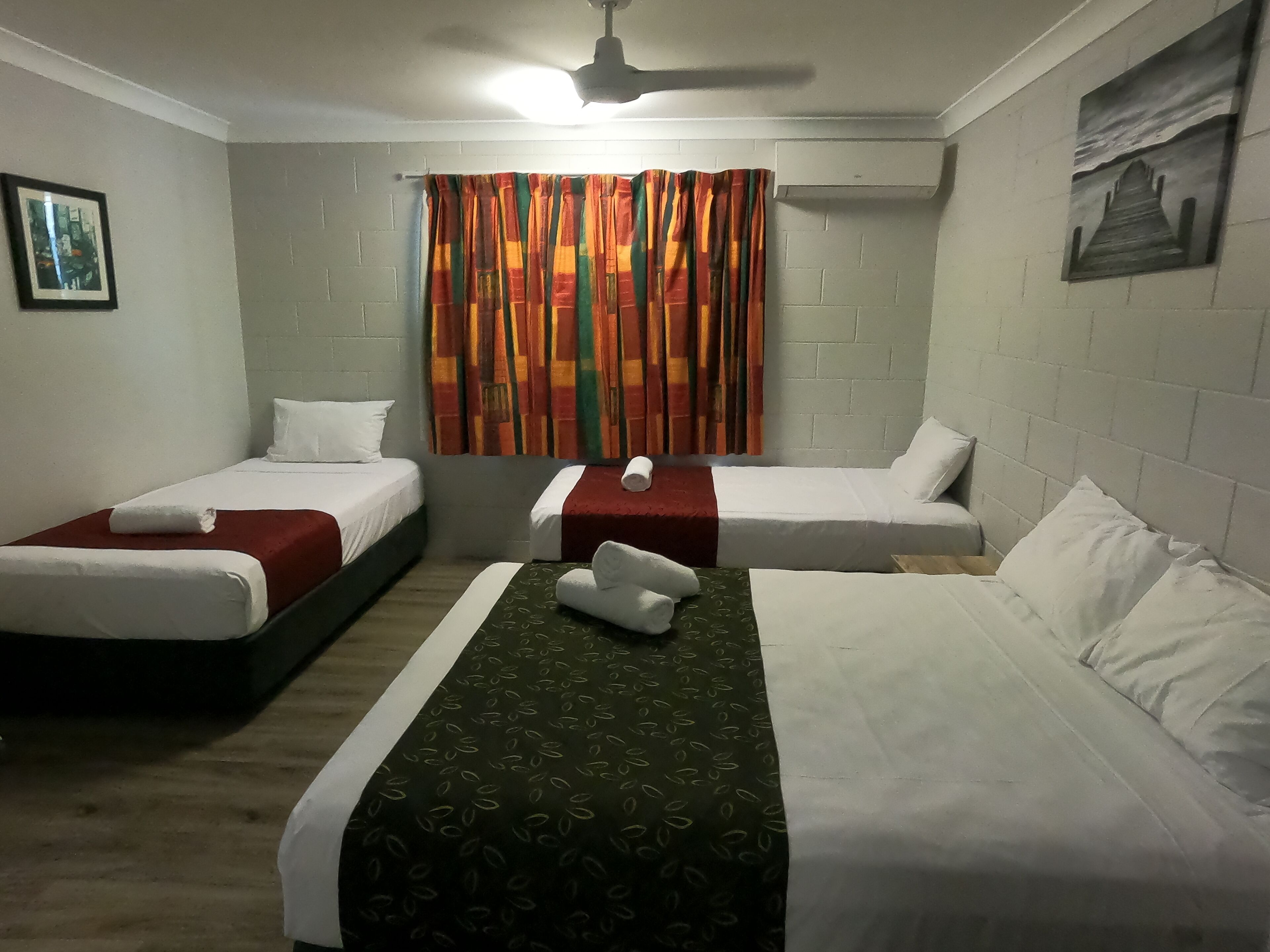 A hotel room with two single beds, a larger bed, a ceiling fan, a window with colorful curtains, and a painting on the wall.