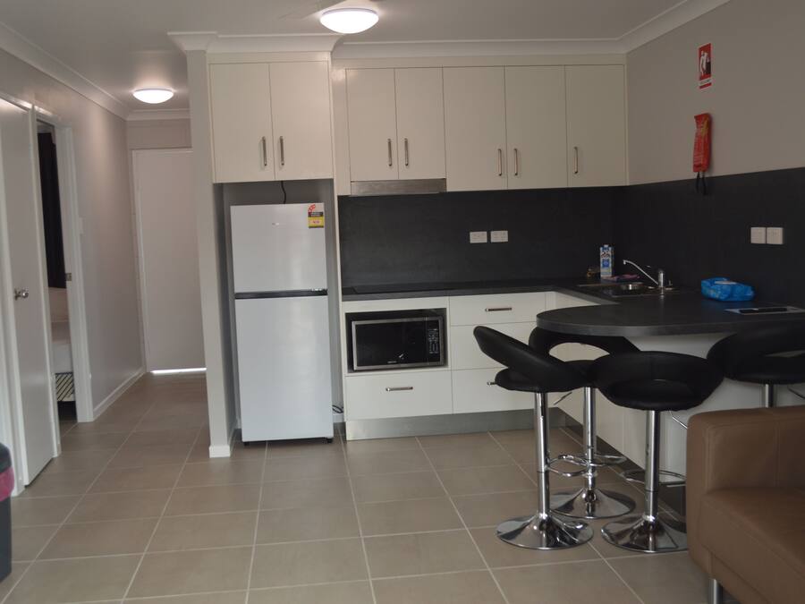 Cozinha moderna com armários brancos, bancada preta e eletrodomésticos de aço inoxidável. Há três banquetas pretas e um sofá marrom.
