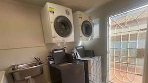 A laundry area with two washing machines, a sink, and a window with a view of a tennis court. - Monte Carlo Motor Inn (Rosslea)