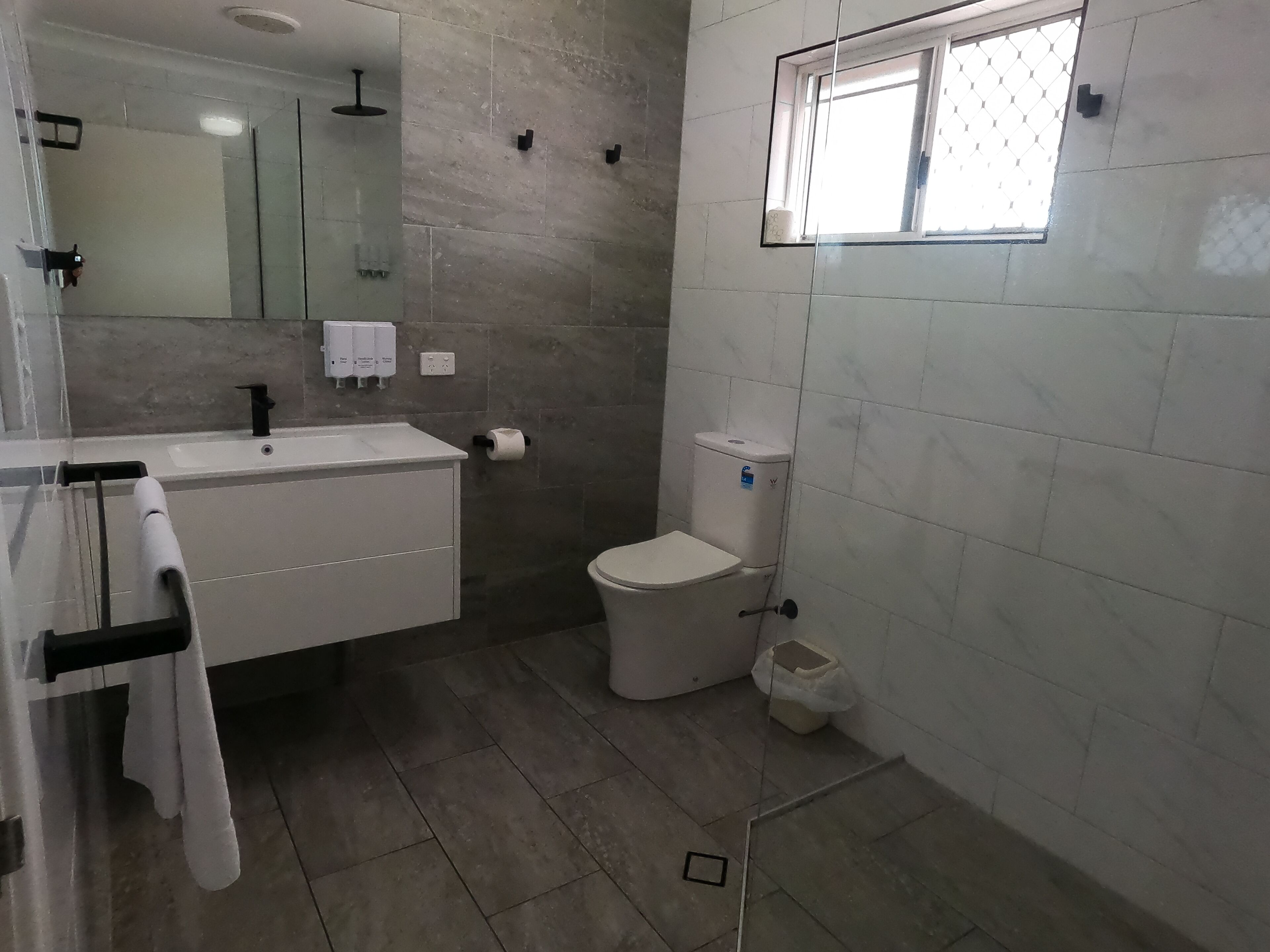 A bathroom with a white sink, mirror, and toilet. There is a window above the toilet and a towel rack with towels.