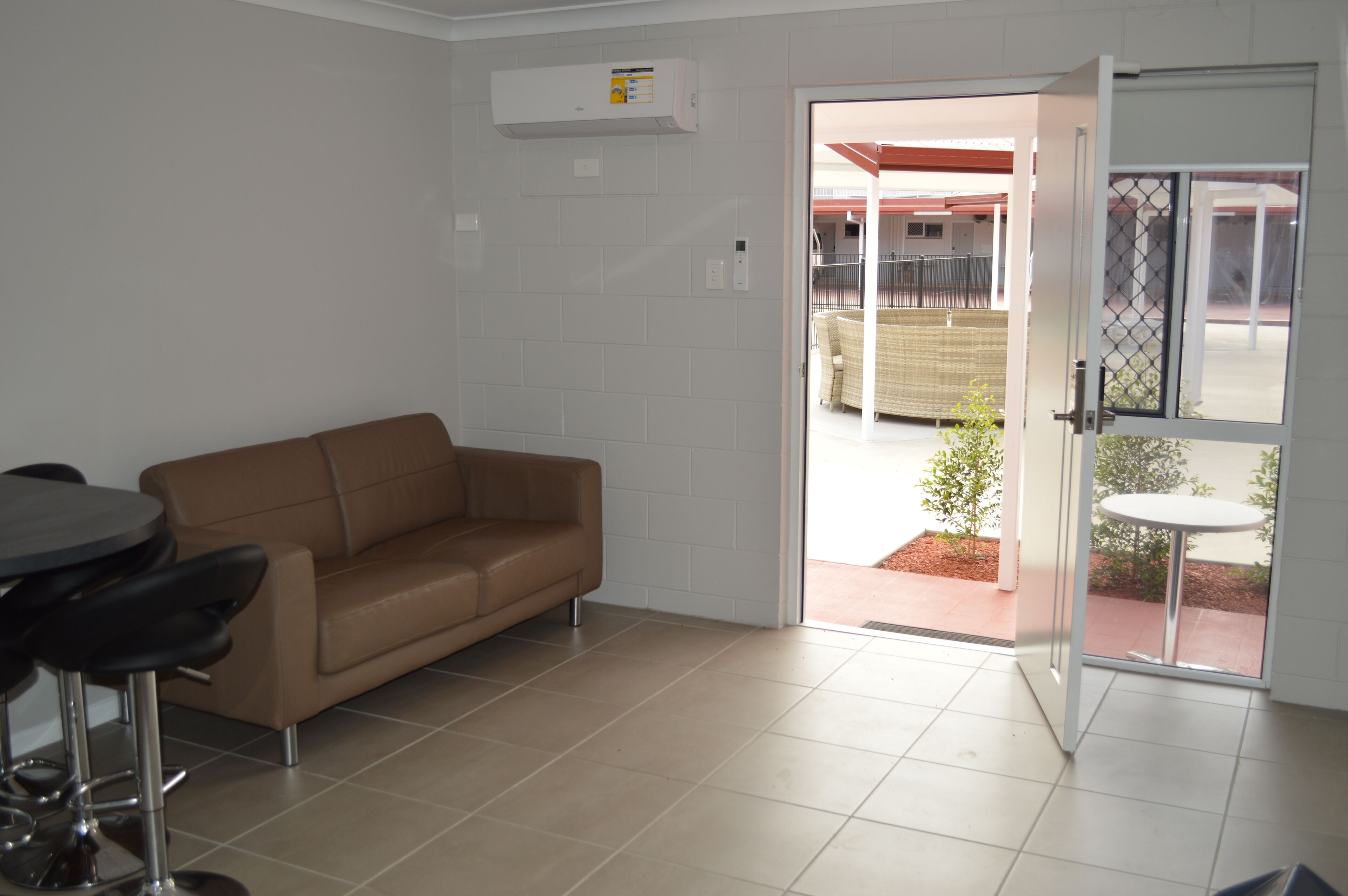 A room with a brown sofa, a round table with chairs, and a sliding glass door leading to an outdoor area with a small table and chairs.