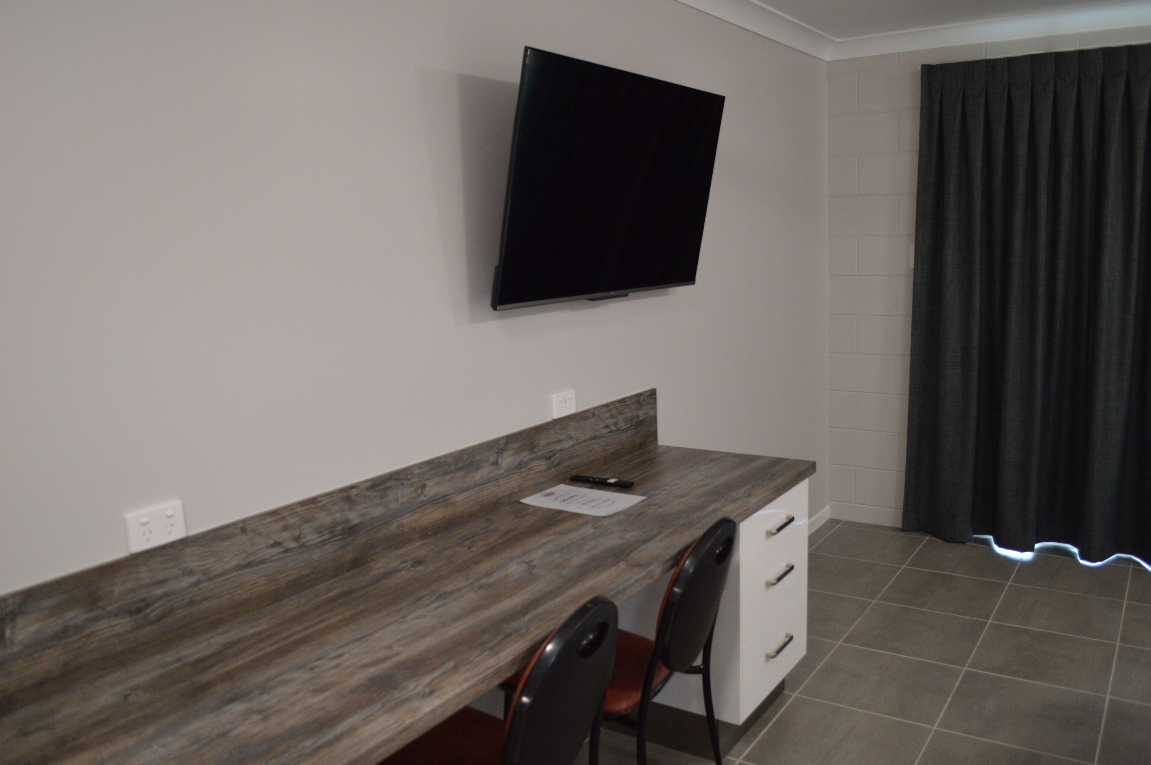 A modern room with a wooden desk, a flat-screen TV mounted on the wall, a window with dark curtains, and a tiled floor.