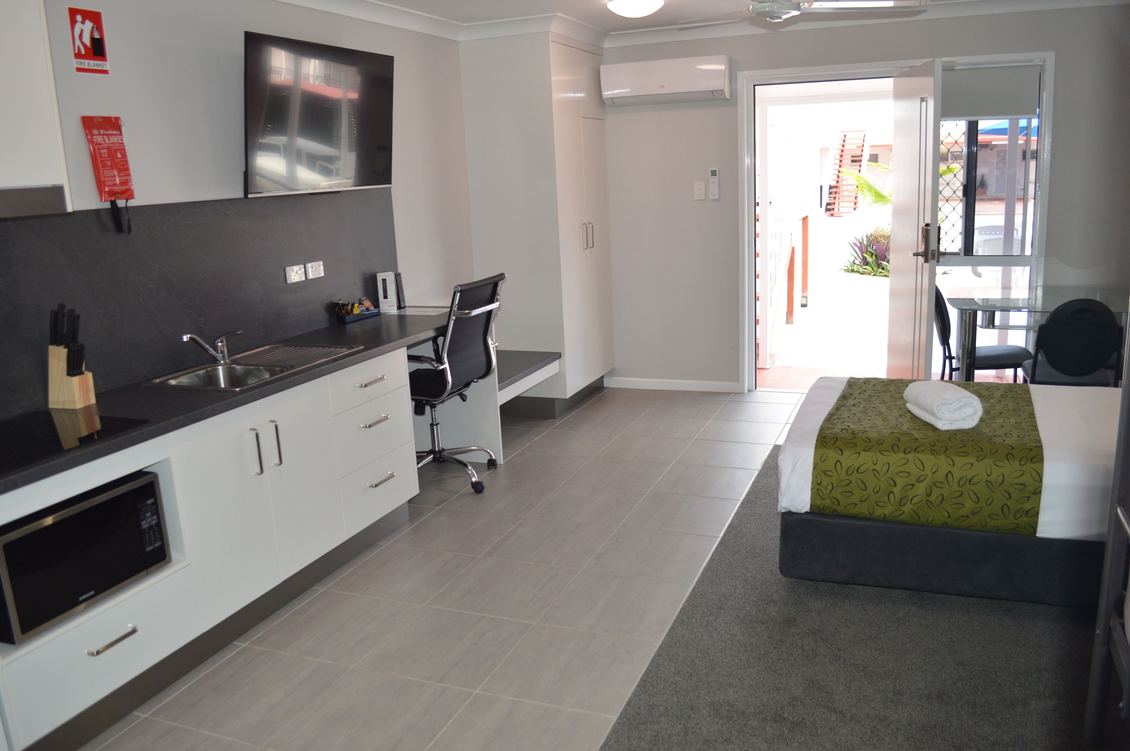 A modern hotel room with a kitchenette, a bed with a green bedspread, and a view of the outside.