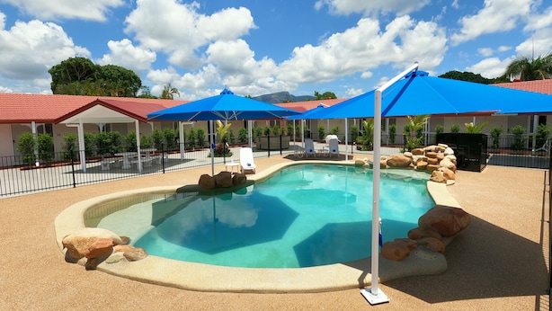 A swimming pool with lounge chairs and umbrellas, surrounded by a fence and a building with a red roof. - Monte Carlo Motor Inn (Rosslea)