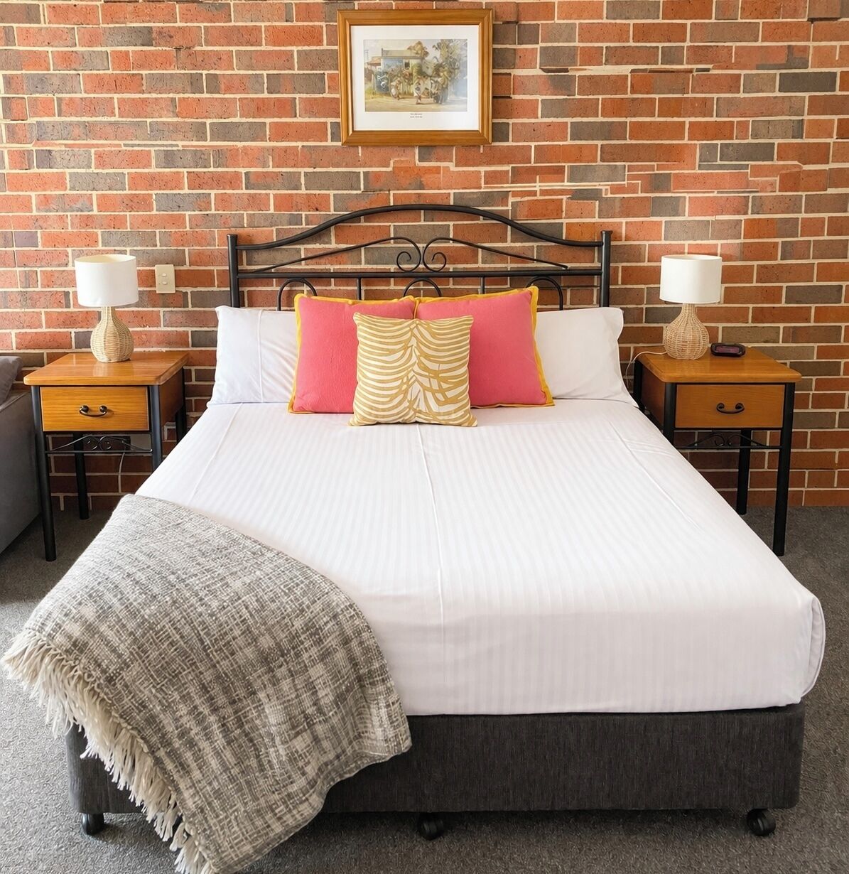 A bed with a headboard, two bedside tables with lamps, a blanket, and a framed picture on a brick wall.