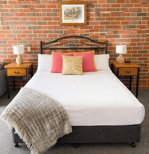 A bed with a headboard, two bedside tables with lamps, a blanket, and a framed picture on a brick wall. - Poachers Paradise Motel (Rutherglen)