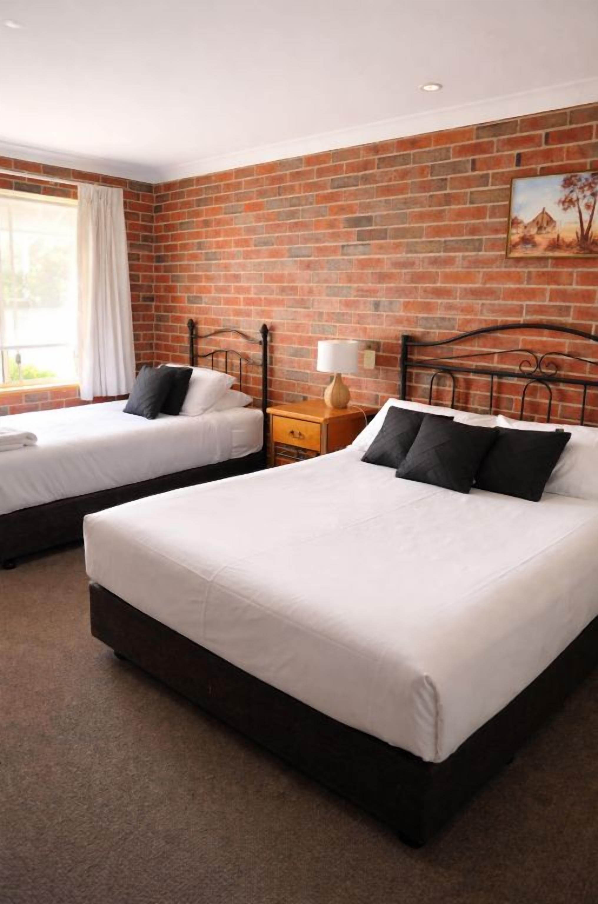 A hotel room with two beds, a brick wall, a window with curtains, and a painting on the wall.