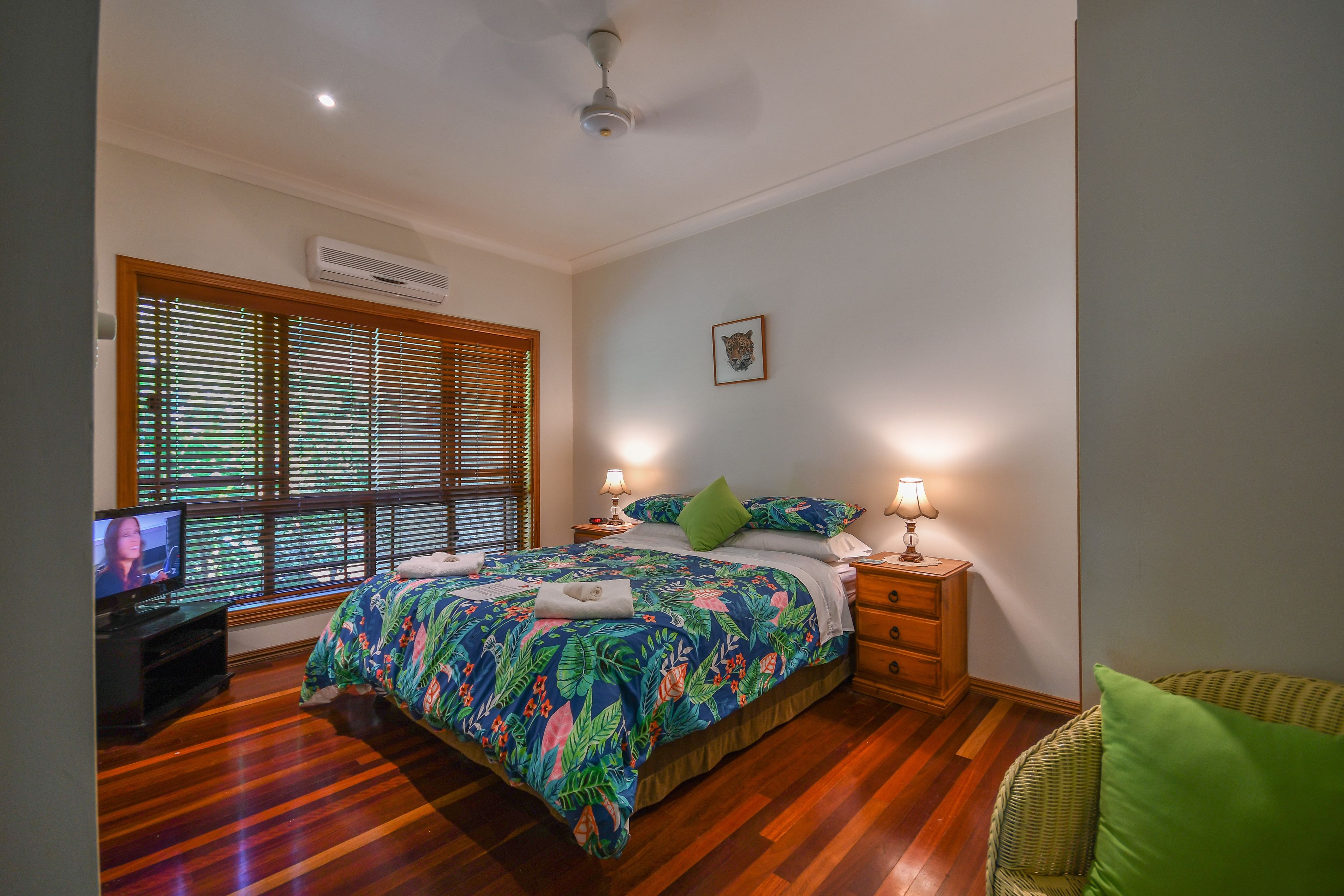 A bedroom with a bed, wooden floor, window with blinds, bedside lamps, and a TV on a stand.