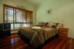 A bedroom with a bed, a nightstand, a lamp, a television, and a window with blinds. - Hibiscus Lodge Bed & Breakfast (Wongaling Beach)