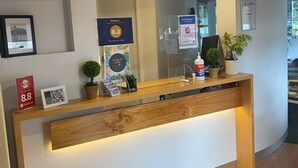 A reception desk with a wooden counter, a mirror above it, and a framed picture on the wall. There are potted plants on the counter and a QR code on the wall. - Fern Bay Motel  (Fern Bay)