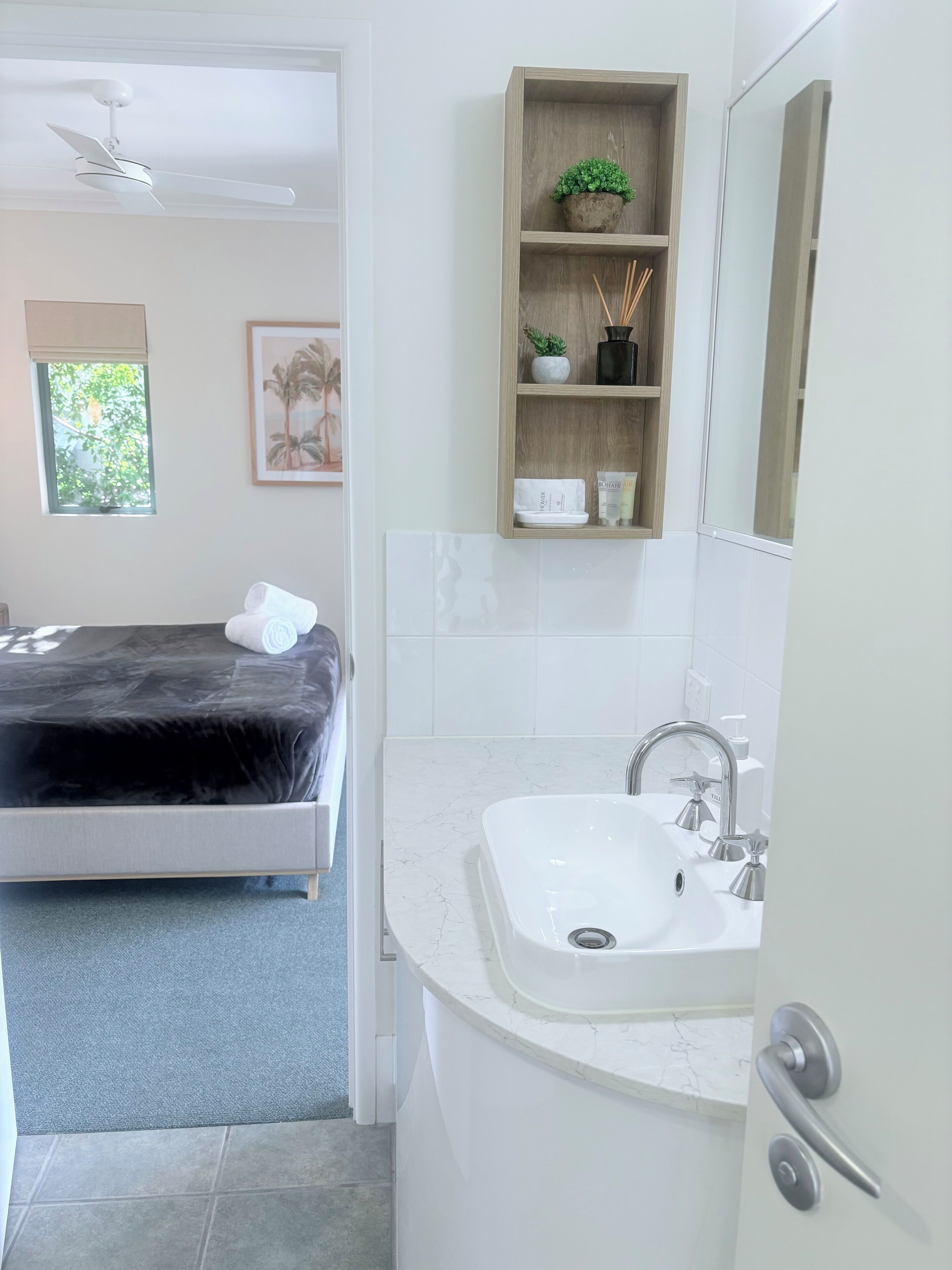 Une salle de bain moderne avec un lavabo blanc, un miroir et une étagère en bois où sont rangés les articles de toilette.