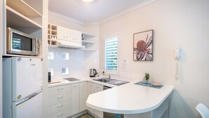 A compact kitchen with white cabinets, a microwave, a refrigerator, and a sink. There is a window with blinds, a framed artwork on the wall, and a phone on the wall. - Noosa River Sandy Shores (Noosaville)