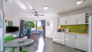 A modern kitchen with white cabinets, a microwave, and a coffee maker. There is a dining area with a round table and green chairs. - Noosa River Sandy Shores (Noosaville)