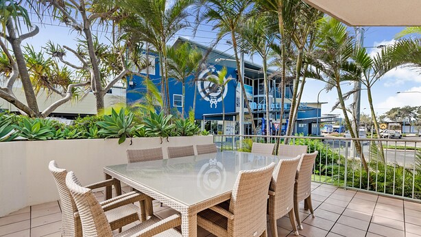 Un espace de restauration en plein air avec une table en verre et des chaises en osier, entouré de palmiers et d’un bâtiment bleu orné d’un logo.