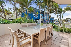 An outdoor dining area with a glass table and wicker chairs, surrounded by palm trees and a blue building with a logo. - Surfcomber on the Beach (Maroochydore)
