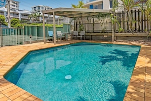 A rectangular swimming pool with a tiled deck, surrounded by a wooden fence and a covered seating area with chairs. - Surfcomber on the Beach (Maroochydore)