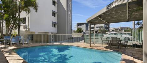 A swimming pool with a covered area, surrounded by a fence, and adjacent to a building with multiple windows and balconies.