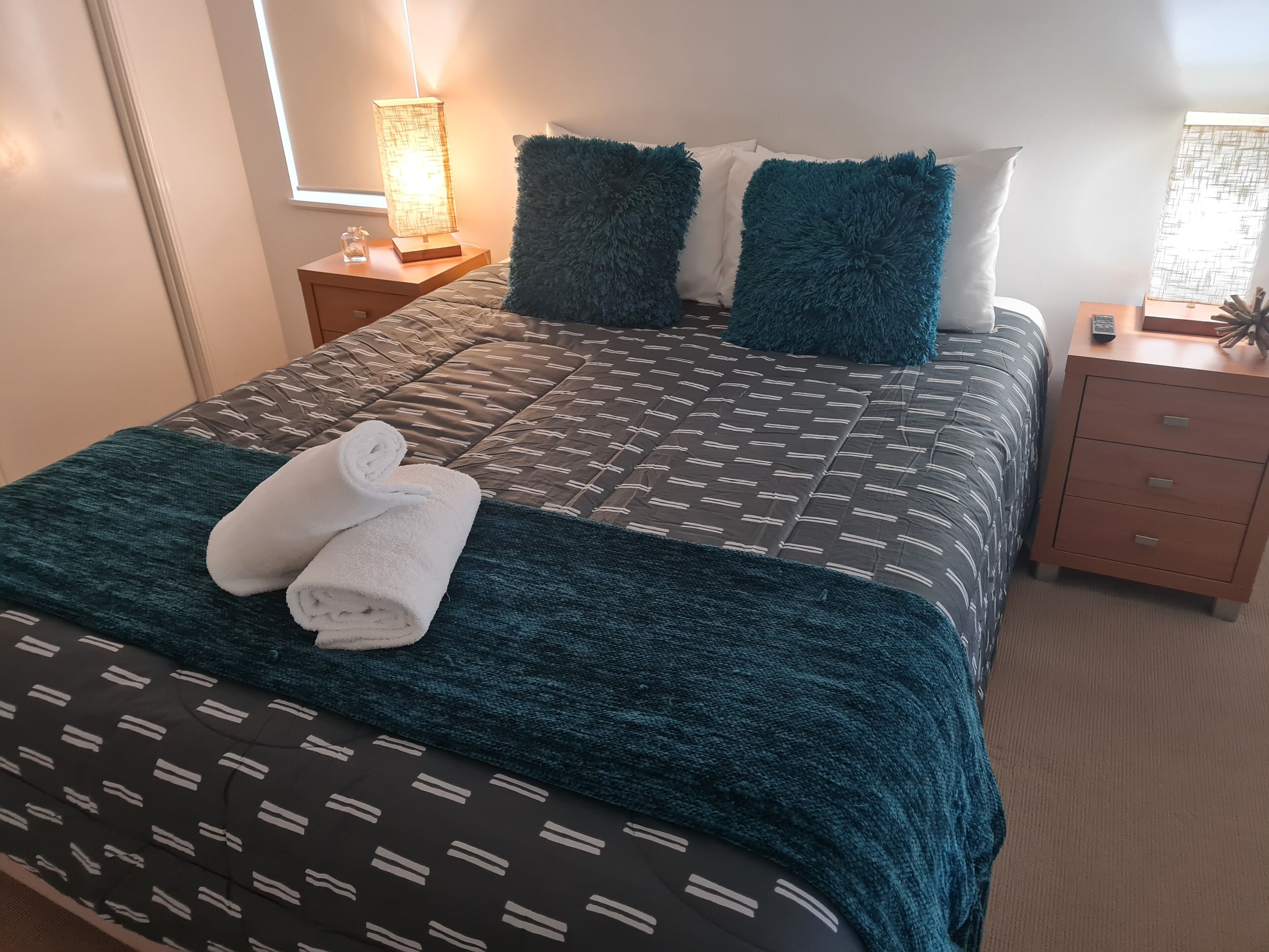 Een opgemaakt bed met een beddegoed met een patroon, twee blauwgroene kussens en een opgerolde witte handdoek op het bed.