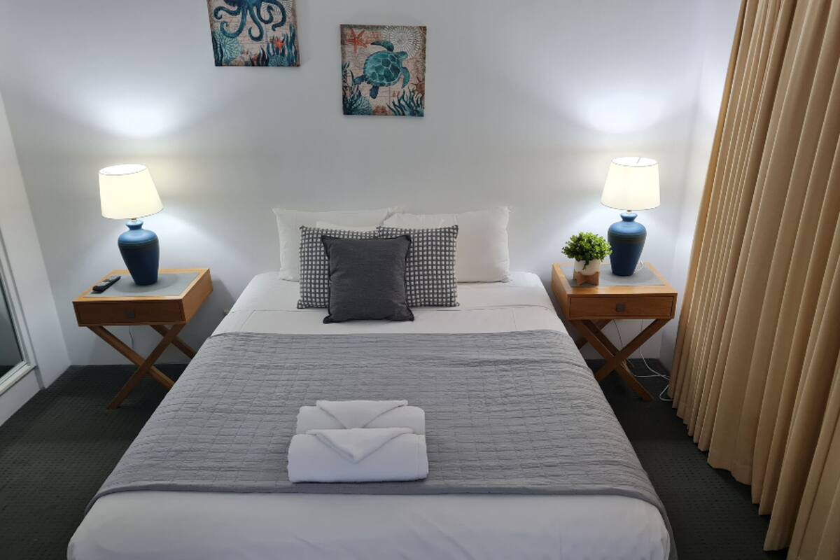 A neatly made bed with a folded towel, two bedside tables with lamps, a small plant, and artwork on the walls.
