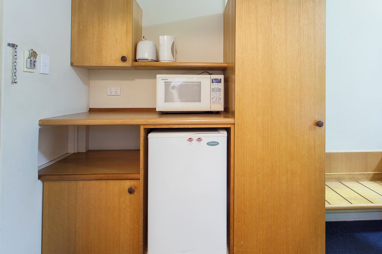 Una kitchenette compacta con microondas, un refrigerador pequeño, armarios de madera y un estante con una tetera y una taza.