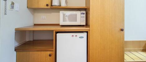 Una kitchenette compacta con microondas, un refrigerador pequeño, armarios de madera y un estante con una tetera y una taza.