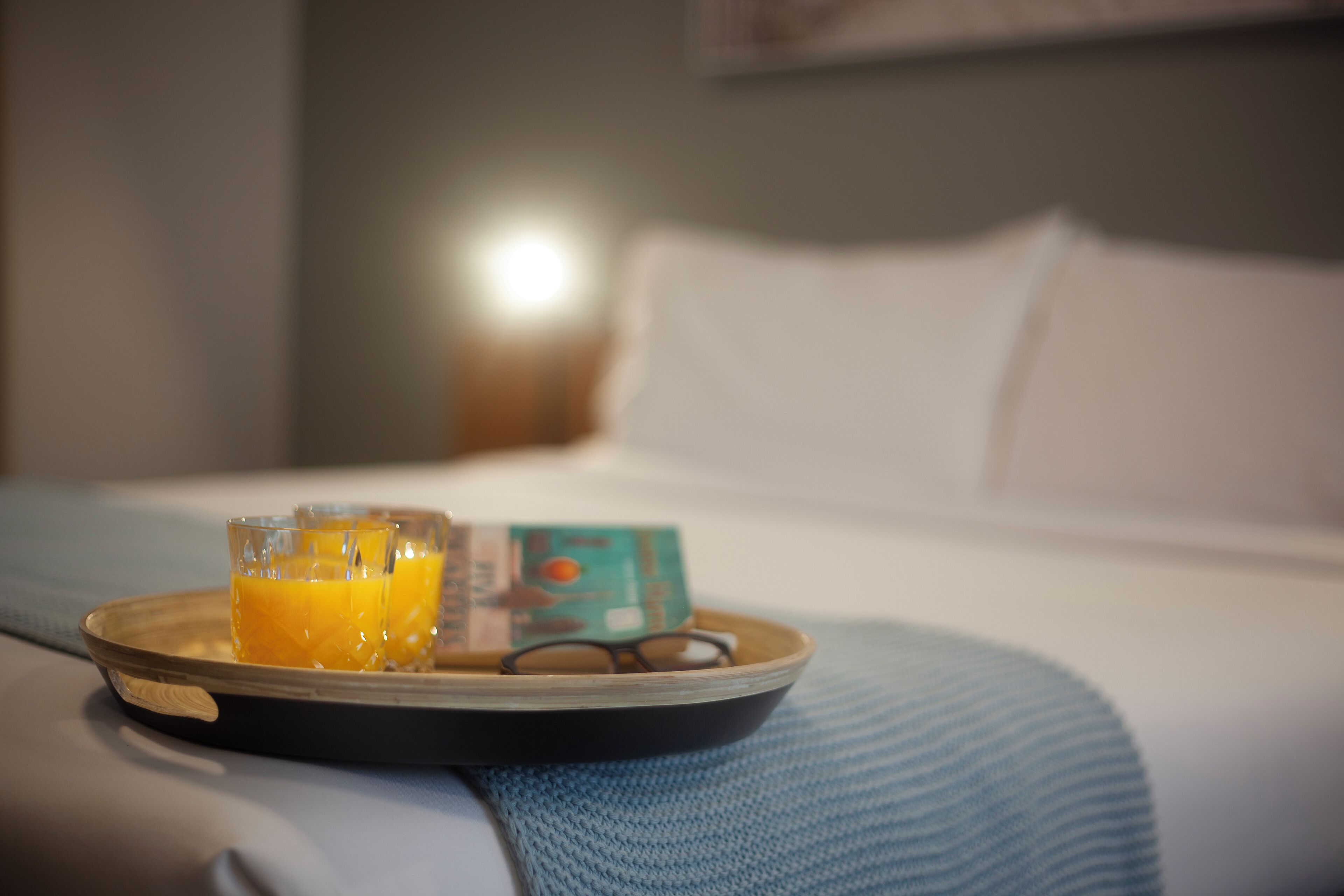 A tray with two glasses of orange juice and a pair of glasses on a bed with a blue blanket.