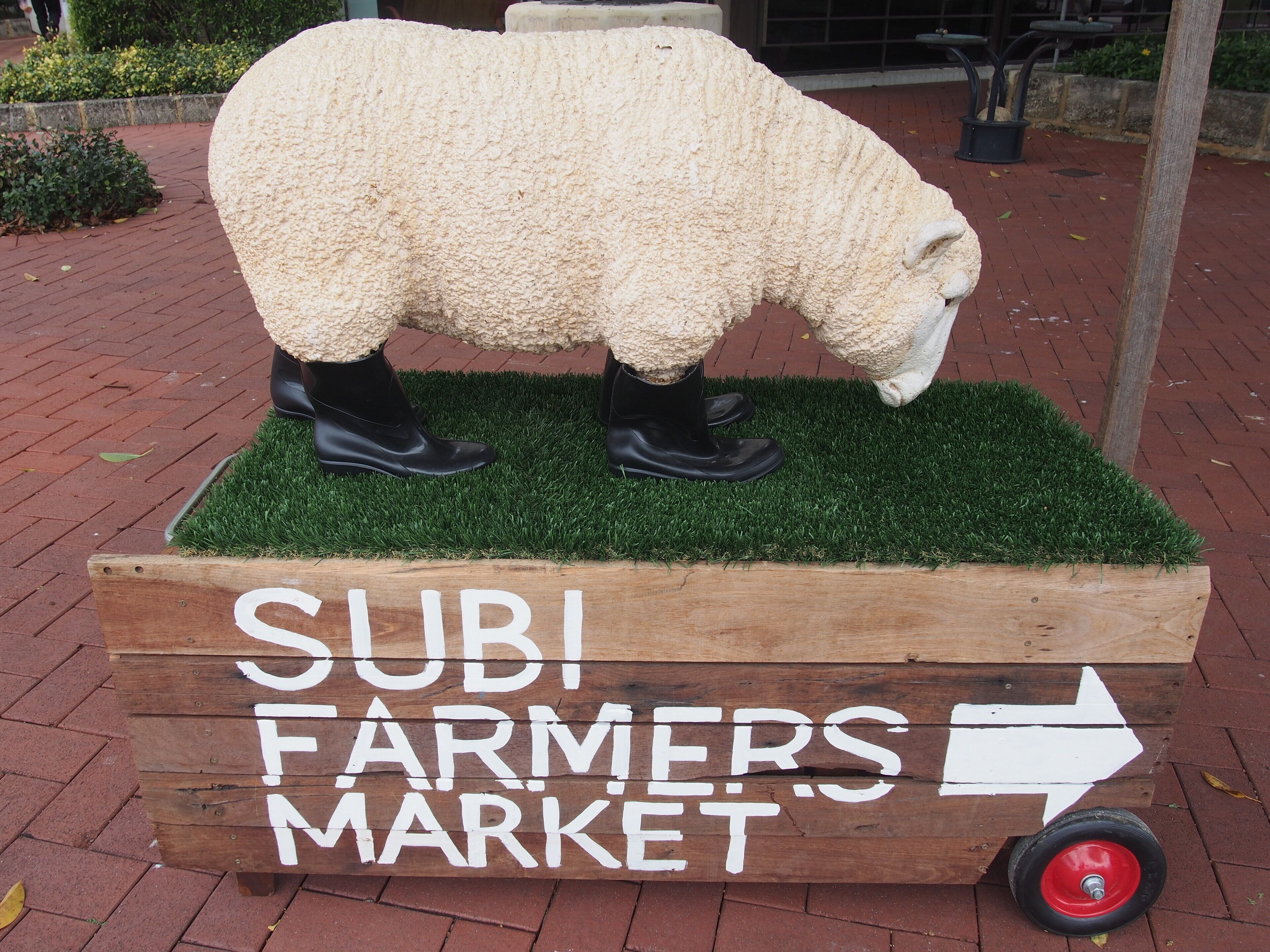 A wooden sign with a sheep sculpture and text indicating a direction to the SUBI FARMERS MARKET.
