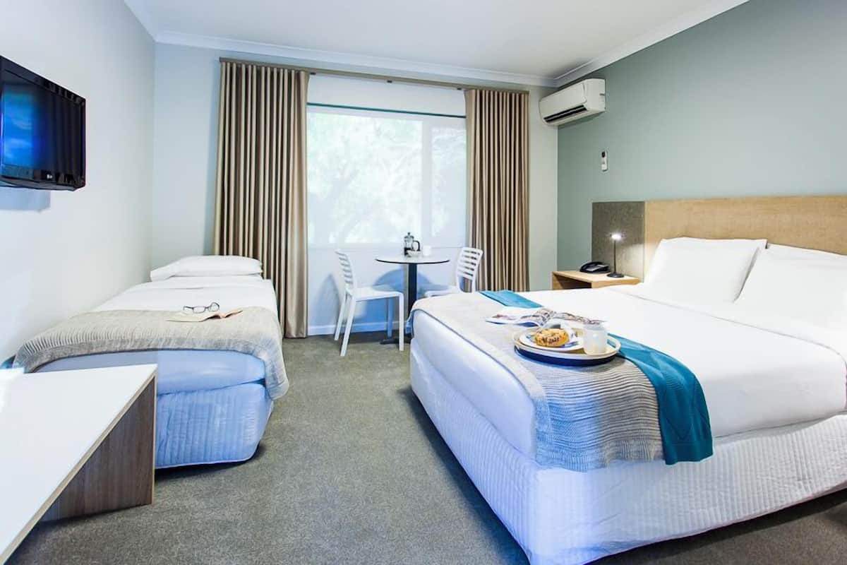 Ein Hotelzimmer mit einem Bett, zwei Einzelbetten, einem Schreibtisch, einem Stuhl, einem kleinen Tisch und einem Fernseher.