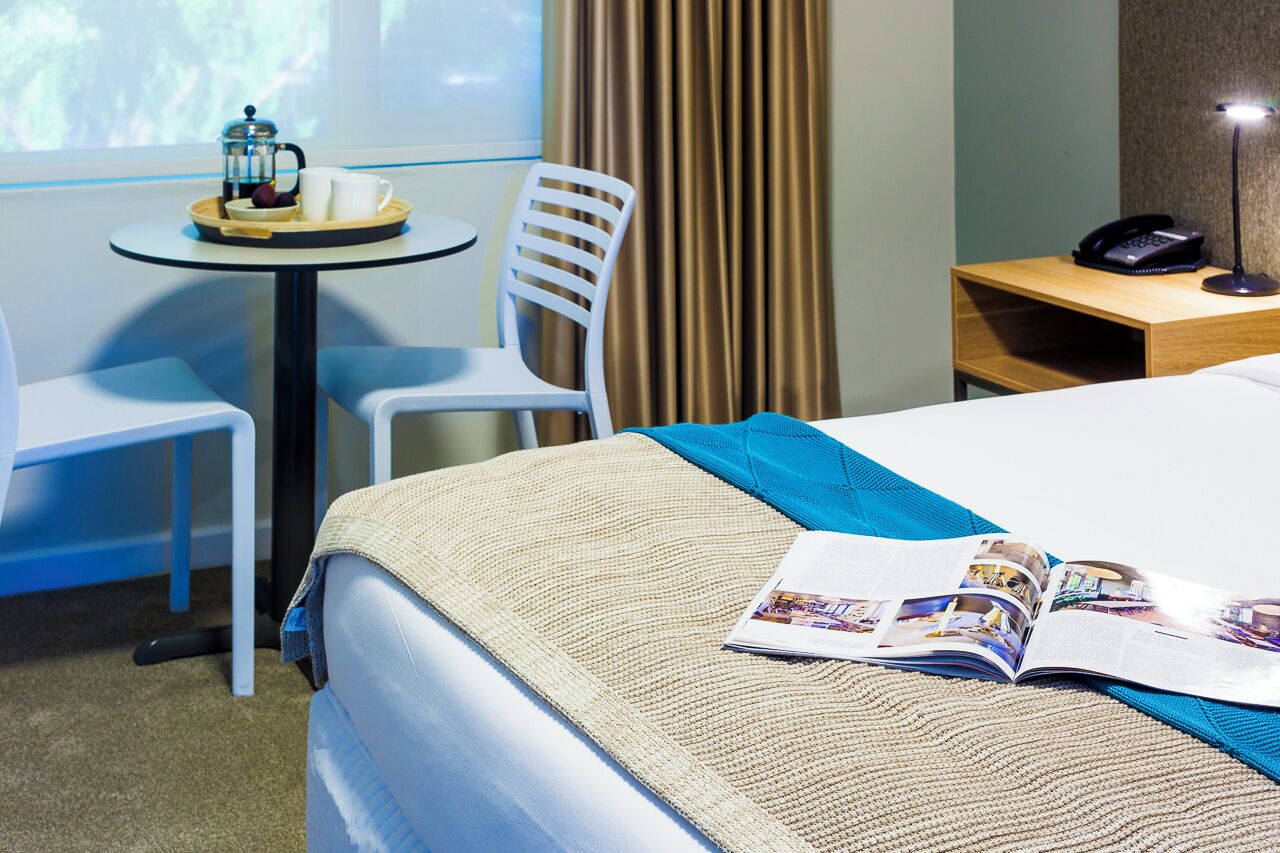 Ein Hotelzimmer mit einem Bett, einem kleinen Tisch mit einer Kaffeemaschine, einem Sessel, einem Nachttisch mit Telefon und einem Fenster mit Vorhängen.