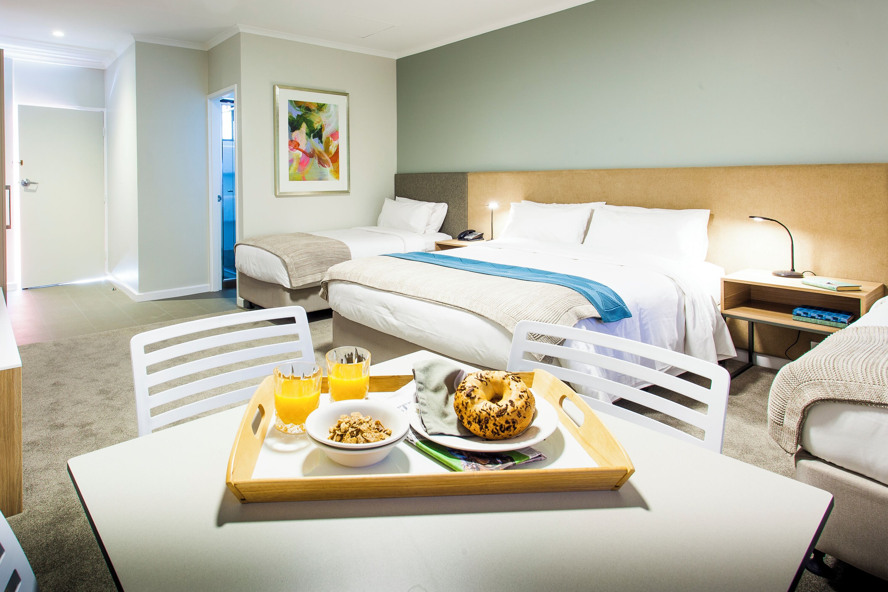 Una habitación de hotel con cama, mesitas de noche, un escritorio con productos para el desayuno y una silla.
