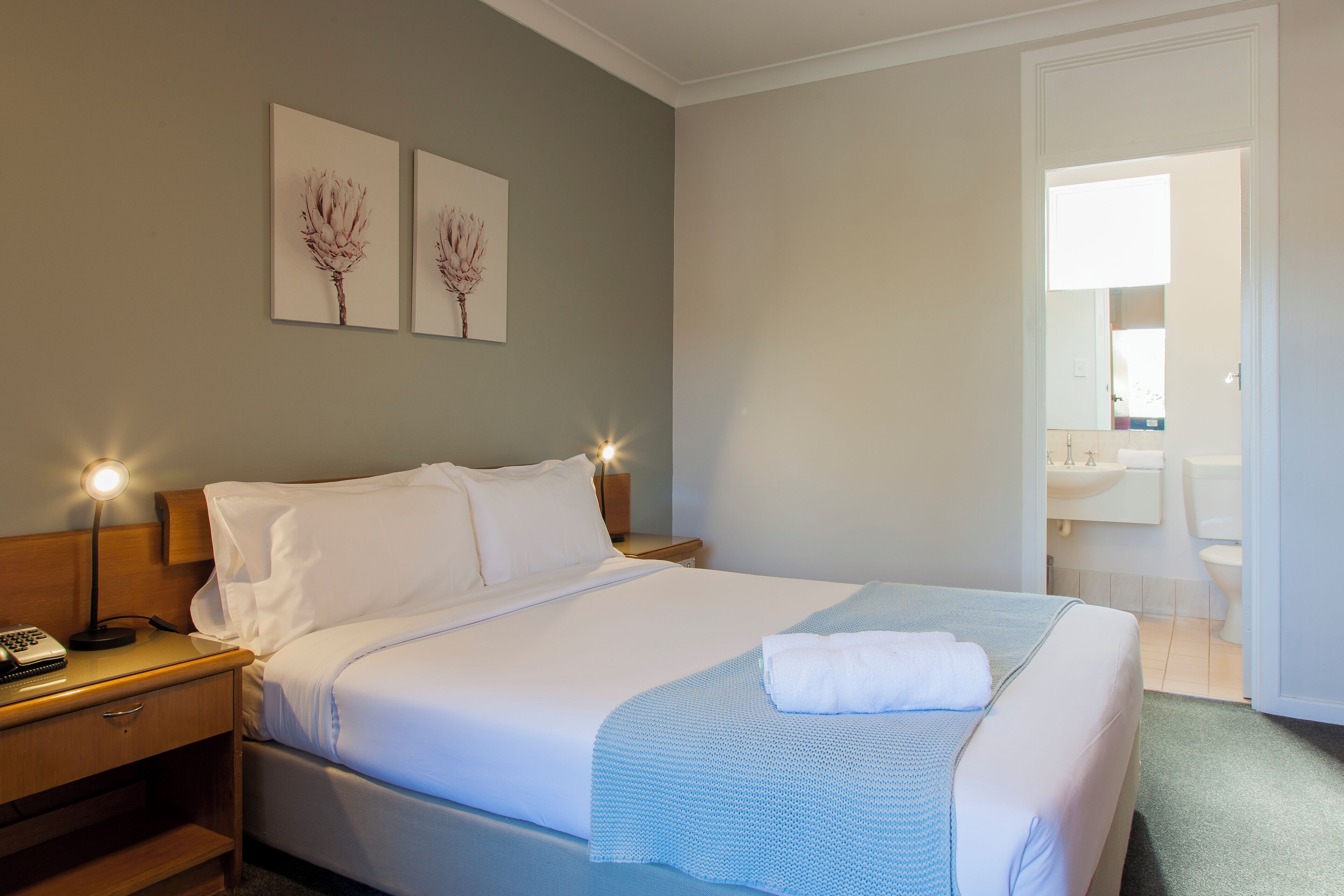 Una cama bien hecha con ropa de cama blanca, una lámpara de noche y dos cuadros en la pared con motivos florales.