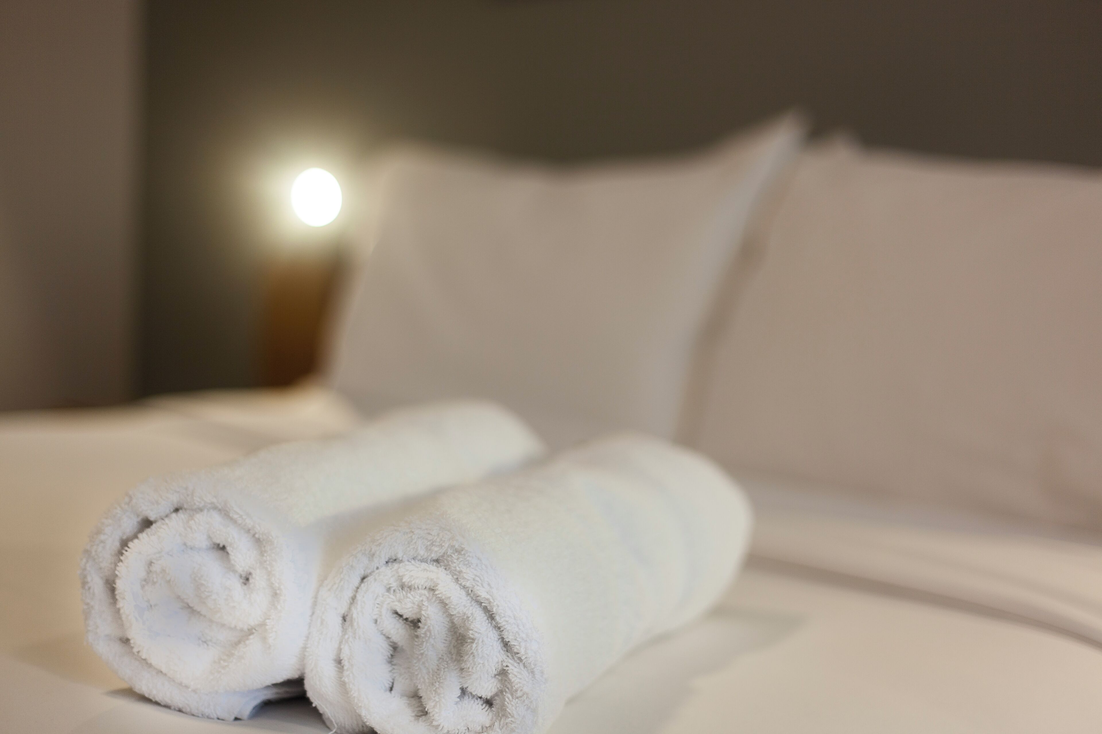 Dos toallas blancas dobladas sobre una cama con ropa blanca y una fuente de luz visible al fondo.