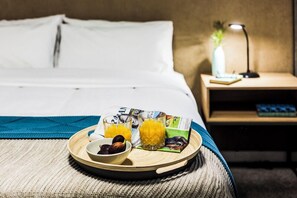 A neatly made bed with white linens, a bedside table with a lamp, a tray with fruit and juice, and a magazine on the bed.
