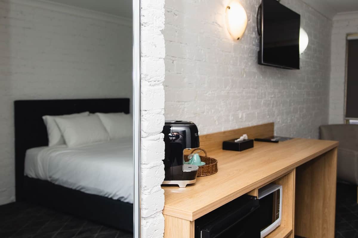 A compact hotel room with a bed, a kitchenette, a flat-screen TV, and a white brick wall.