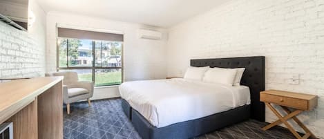 Un dormitorio con cama, escritorio y silla. Se ve follaje verde a través de la ventana.