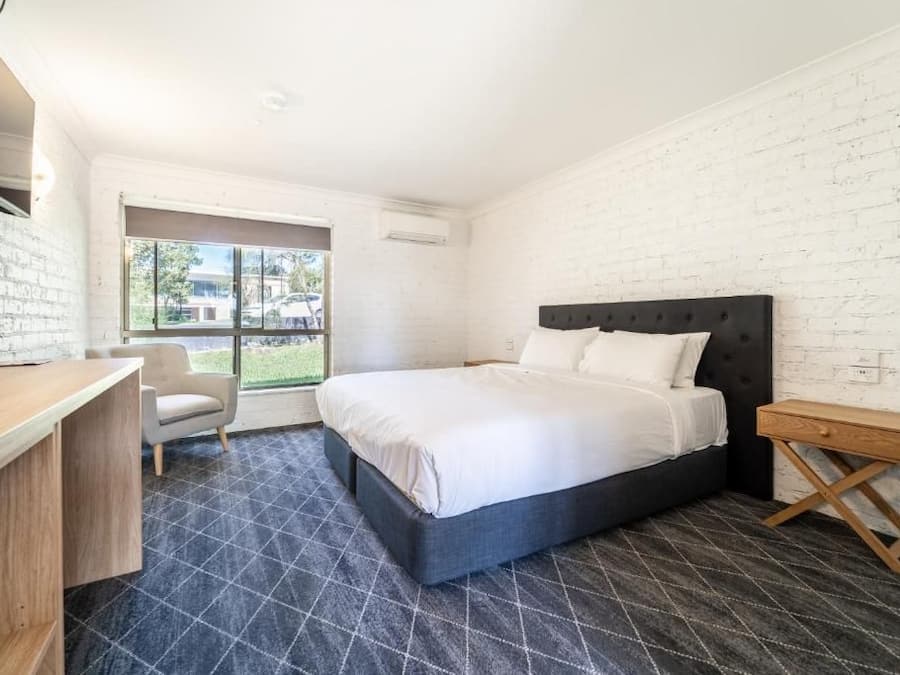 Un dormitorio con cama, escritorio y silla. Se ve follaje verde a través de la ventana.