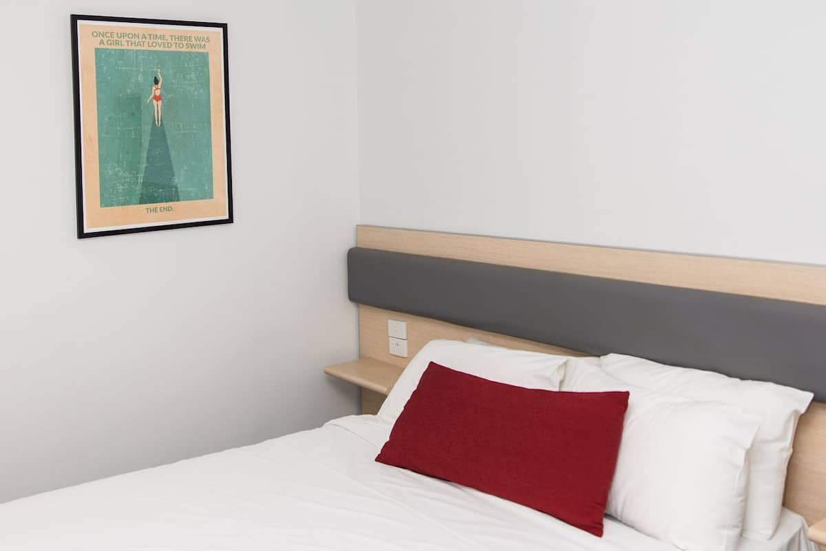 A bedroom with a bed, a nightstand, a red pillow, and a framed poster on the wall.